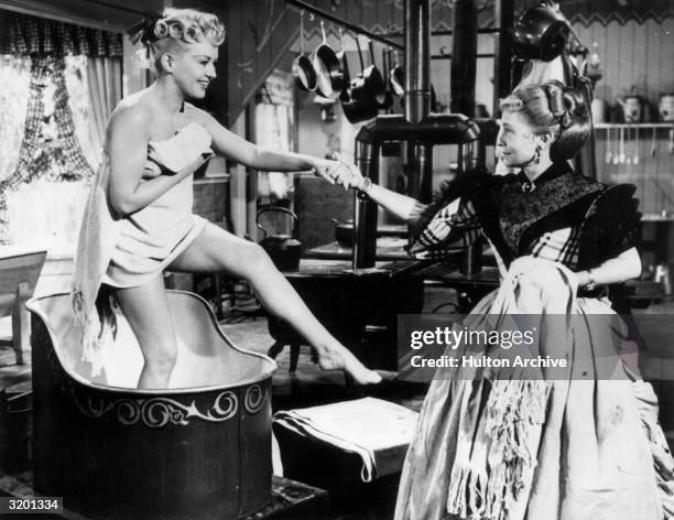 American actor Betty Grable steps out of a wash basin wearing a towel, while Thelma Ritter holds her hand to give her support, in a still from...