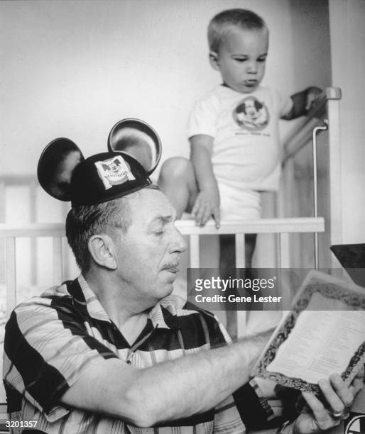 American cartoonist and movie studio head, Walt Disney , wearing Mickey Mouse ears, reads his grandson a story from a children's book. His grandson...