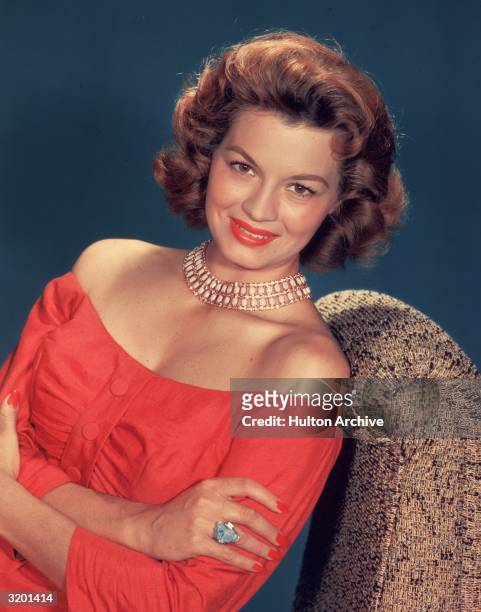 Portrait of American actor Angie Dickinson seated, wearing a coral, off-the-shoulder dress. She wears an ivory, jeweled necklace.