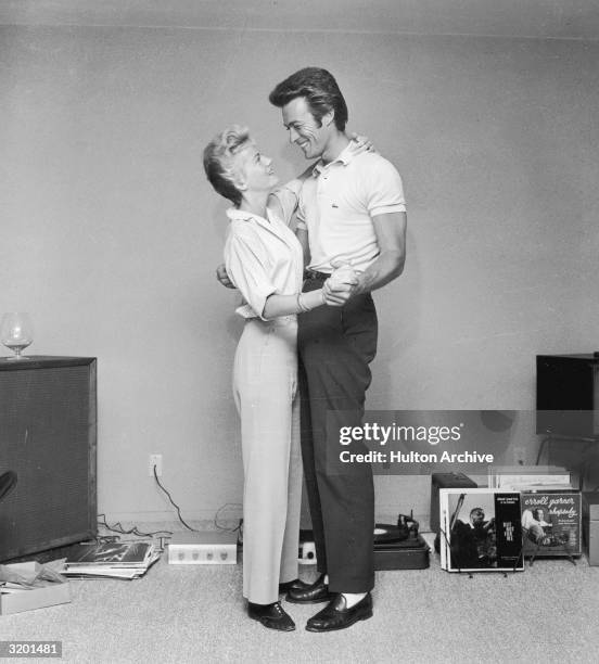 Full-length image of American actor Clint Eastwood and his first wife, Maggie, dancing next to a turntable and a rack full of records in a living...
