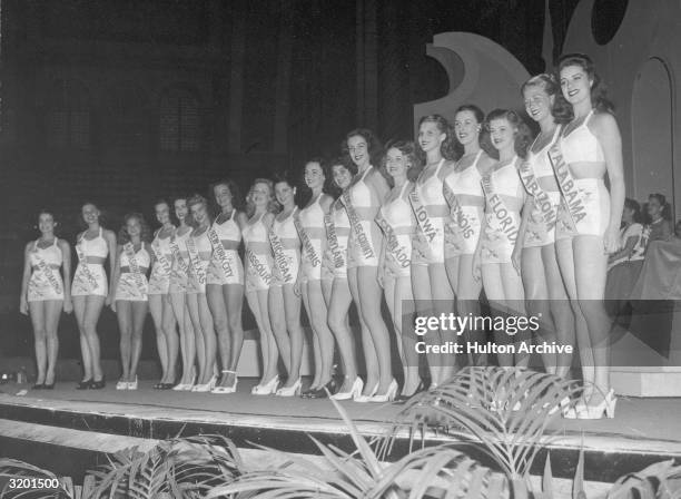 Contestants in the Miss America pageant smile, as they stand onstage in a row, wearing swimsuits and sashes representing their home states.