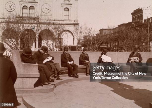 Full-length view of men, some reading newspapers, seated on a circular granite bench at Herald Square, near 34th Street, New York City.