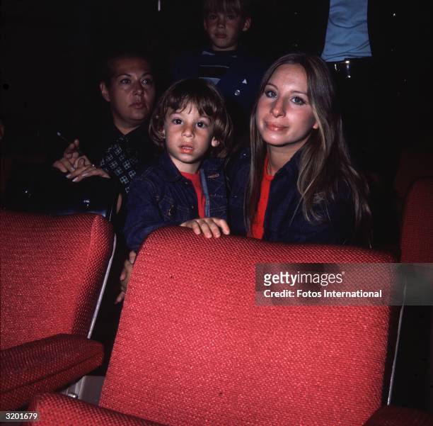 American singer and actor Barbra Streisand, seated behind a red movie theater seat, with her young son Jason and unidentified theater patrons. Jason,...