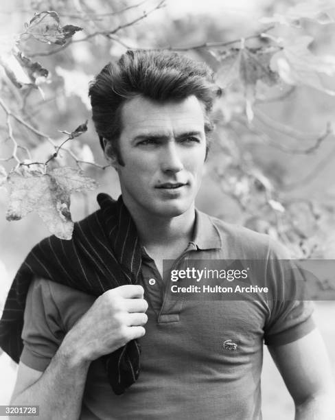 Portrait of American actor and director Clint Eastwood squinting while posing under a tree, with a sweater slung over his shoulder. Eastwood wears a...