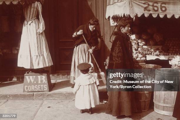 Full-length image of a woman and two young children standing on a sidewalk between a produce store and a corset maker's shop, while another woman...