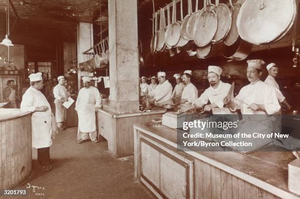 Full-length view of cooks, wearing aprons and chef hats, preparing food behind two long counters in the kitchen of Delmonico's restaurant at the...