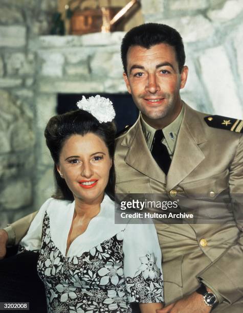 Portrait of American actor Barbara Stanwyck and her second husband, actor Robert Taylor, smiling while posing on a sofa. Stanwyck wears a floral...