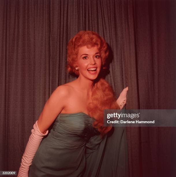 Portrait of American actor Donna Douglas, wearing a blue shoulderless gown and long white gloves, standing in front of a curtain and smiling.