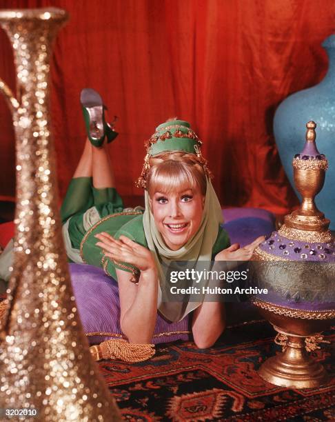 American actor Barbara Eden holds her hands up and smiles while lying on her stomach on top of cushions on an oriental rug, in a promotional portrait...