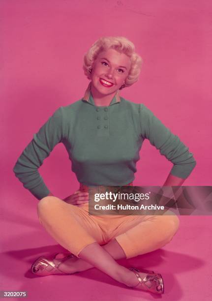 Full-length studio portrait of American actor and singer Doris Day sitting on the floor in front of a pink backdrop, her legs crossed and her hands...