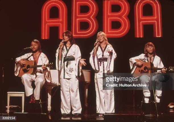 Swedish pop group ABBA , wearing similar white Asian-influenced costumes, perform for the television program Midnight Special with a neon 'ABBA' sign...