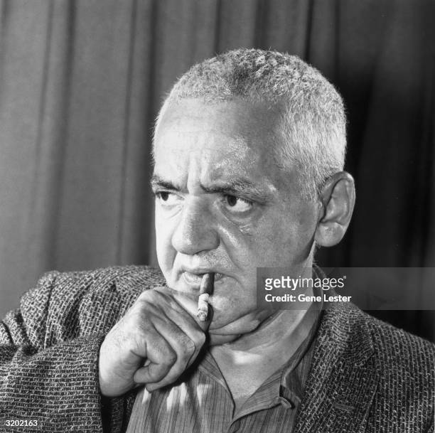 Portrait of American photojournalist Arthur 'Weegee' Fellig gripping his chin, while a cigar dangles between his lips.
