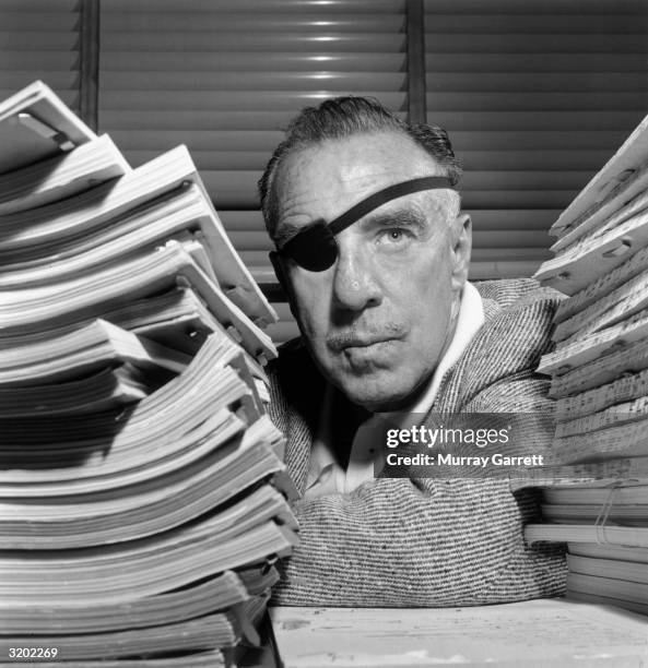 Headshot portrait of American film director Raoul Walsh leaning on a table between two stacks of film scripts in his office at Warner Brothers...