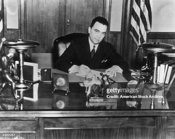 Portrait of FBI director J. Edgar Hoover sitting at a desk while signing a document.