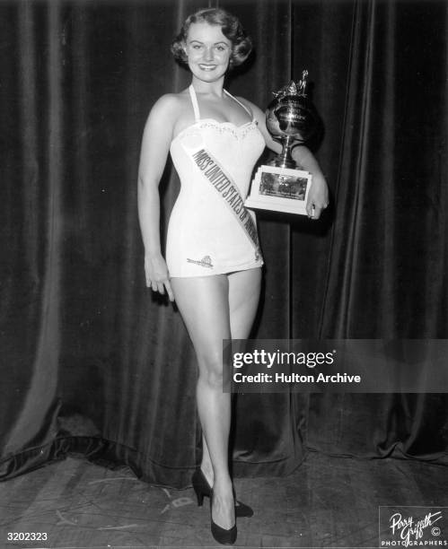 Full-length portrait of Myrna Hansen, Miss United States of America and second runner-up in the Miss Universe Pageant, holding a trophy while wearing...