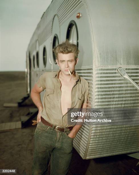 American actor James Dean leaning against a dressing room trailer with his shirt open to the waist while smoking a cigarette on the set of director...