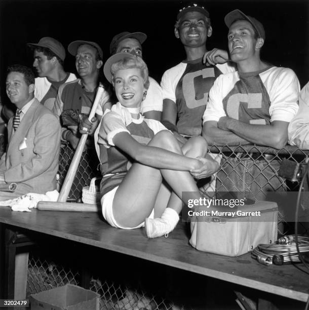 American actor Janet Leigh sits on a dugout bench while her teammates, actor Vince Edwards, jazz trumpeter Harry James, and actors Tony Dante, Jeff...