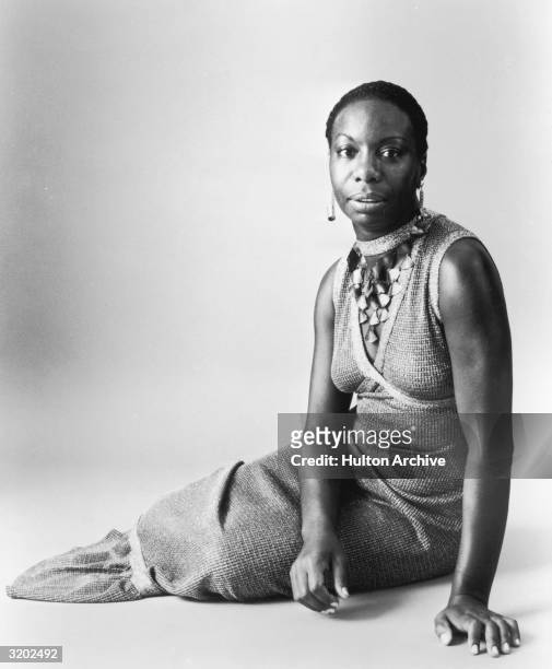 Studio portrait of American pianist and jazz singer Nina Simone reclining on the floor in a sleeveless, V-neck dress with a shell neckpiece.