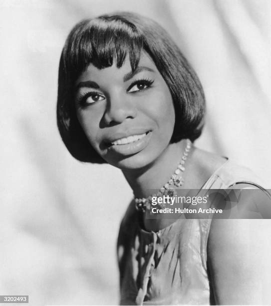 Headshot portrait of American pianist and jazz singer Nina Simone smiling over her shoulder. Simone wears a sleeveless dress and a choker.
