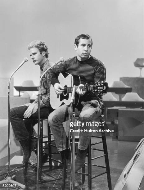 American folk rock duo Simon and Garfunkel performing on a studio sound stage during a rehearsal. Garfunkel slaps his leg while Simon plays a guitar.