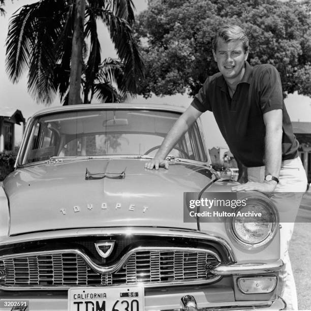 Portrait of American actor Troy Donahue leaning on the hood of a Toyota Toyopet car with a California license plate, in front of a palm tree on a...