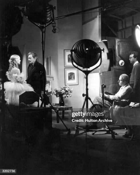 American film director Howard Hawks sits in a folding chair and watches American actors Carole Lombard and John Barrymore perform a scene on the set...