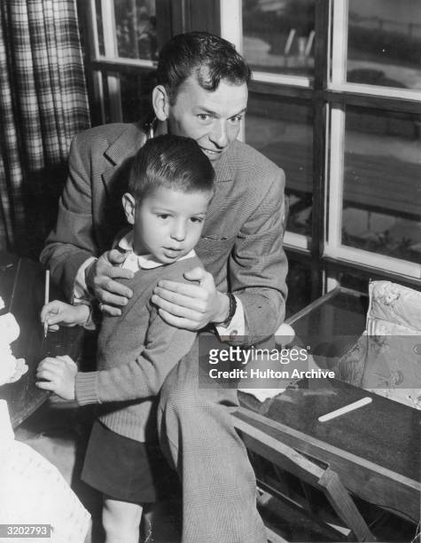 American singer and actor Frank Sinatra smiles while holding his young son, Frank Jr., near a window.