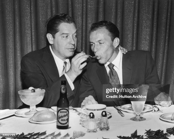 American comedian Milton Berle feeds singer and actor Frank Sinatra with a spoon while sitting with fruit cocktail cups in front of them at a banquet...