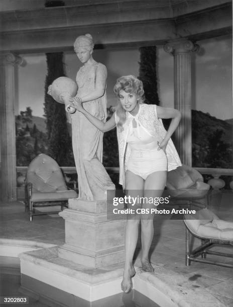 American actor Donna Douglas poses in a bathing suit while dipping her toe into a swimming pool on the set of the television series, 'The Beverly...