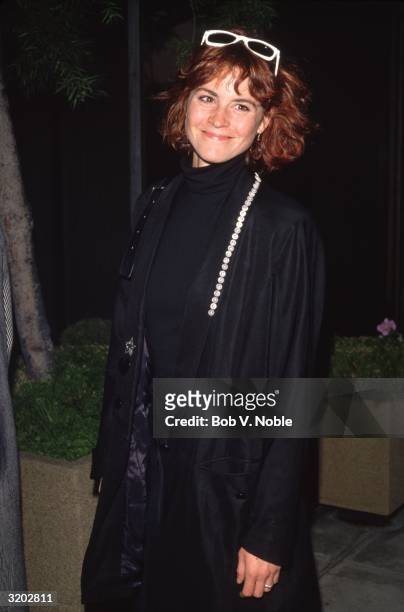 American actor Ally Sheedy smiles while wearing a long black coat and white-framed sunglasses on top of her head.