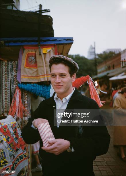 American actor Paul Newman cradles a soda bottle under one arm while digging into a small snack food bag in front of a stall selling Mexican...