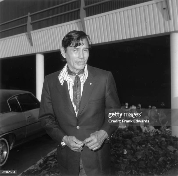 Native American actor Jay Silverheels standing outside a garage. Silverheels is wearing a sport coat over a wide-collar shirt and Western tie.