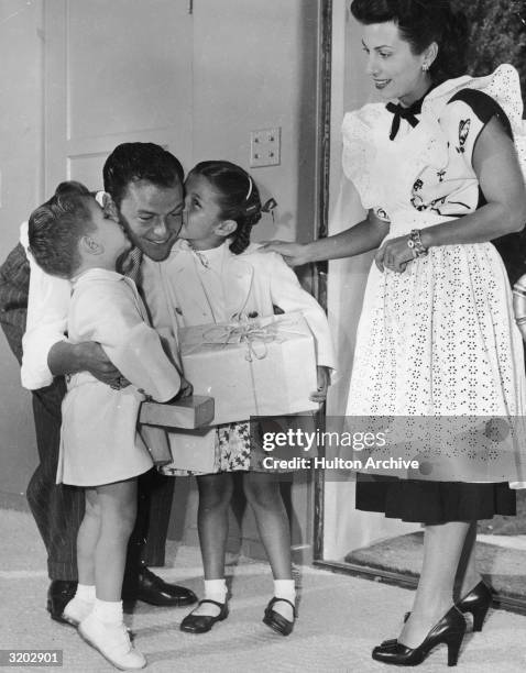 American singer Frank Sinatra bends down to be welcomed home with a kiss on either cheek from his children, Frank Jr. And Nancy Jr., while his wife,...