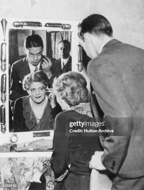 American actor Ethel Barrymore sits in front of a mirror at a dressing table, applying a makeup stick to her eyebrow while actor John Drew Colt...