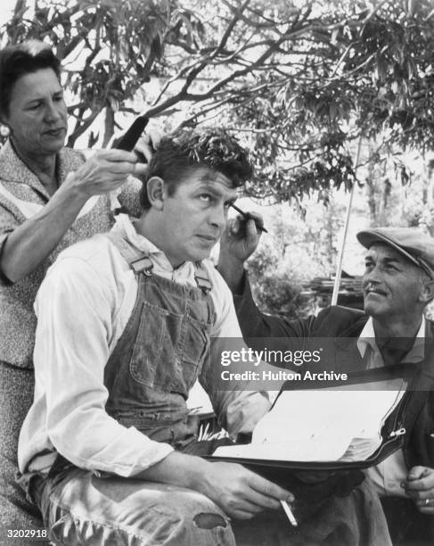 American actor Andy Griffith, wearing overalls and a work shirt, sits as hairdresser Buddy King and makeup artist Newt Jones prepare him for a scene...