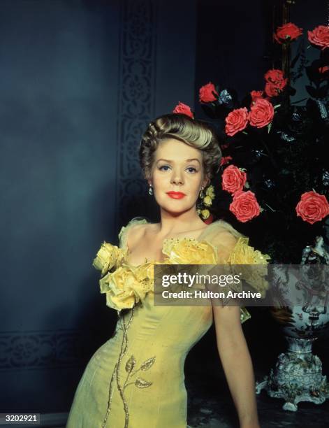 American singer and actor Alice Faye wears a yellow velvet saloon performer's dress with roses at the neckline while posing beside an arrangement of...