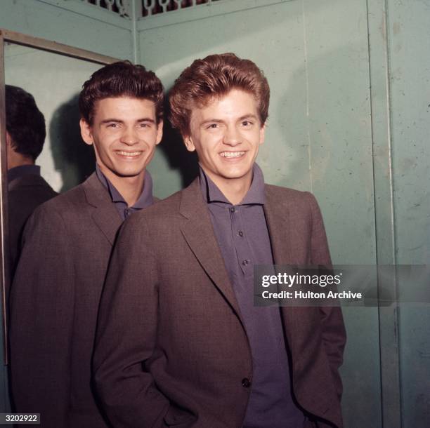 Portrait of the American rock and roll group The Everly Brothers smiling while posing with their backs to a mirror in polo shirts and jackets, 1960s.