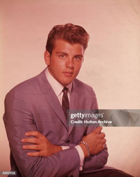 Studio portrait of American singer and actor Fabian seated with his arms crossed in front of his chest while wearing a gray suit and striped tie,...