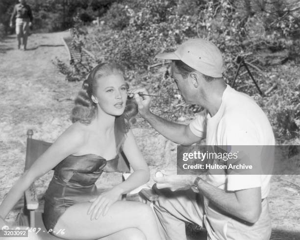 American actor Joan Caulfield sits in a folding chair while a makeup artist prepares to use a fine brush on her eyes on the set of director Henry...