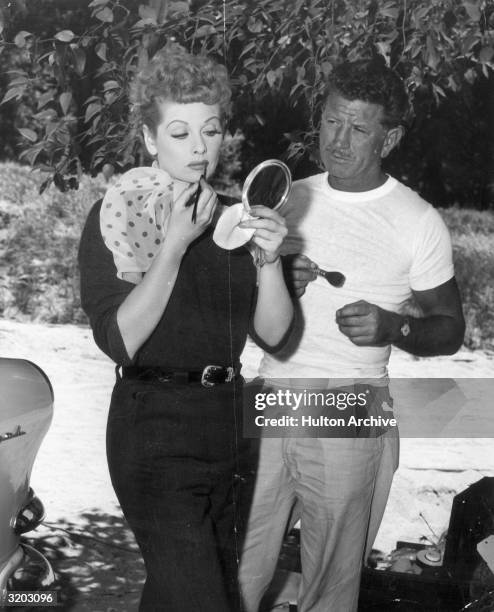 American actor Lucille Ball looks into a handheld mirror while applying lipstick with a brush in front of a makeup artist. A makeup case sits in the...