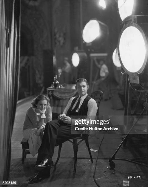 American actor Joan Crawford sits on a stool with her face in her hand while Clark Gable smokes a cigarette in a chair next to her on the set of...