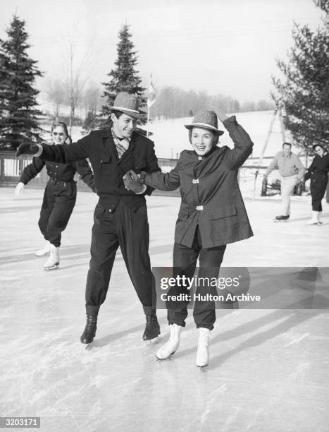 Full-length image of married American singer/actors Eddie Fisher and Debbie Reynolds ice skating on an outdoor rink. Both wear matching hats.