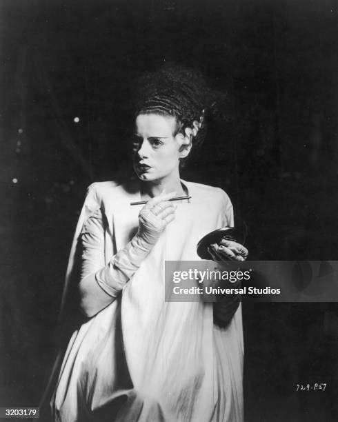 British actor Elsa Lanchester stands in a spotlight surrounded by darkness while holding an eyebrow pencil and a compact mirror on the set of...