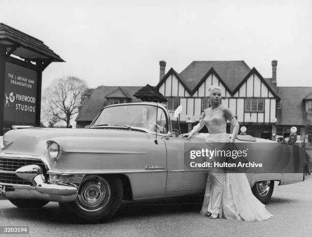 Full-length portrait of British actor Diana Dors posing next to a Cadillac convertible at the entrance gates of Pinewood Studios, Iver Heath,...