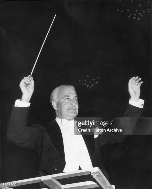 American classical conductor Arthur Fiedler raises his baton while standing at a podium during a concert. Fiedler wears white tie and tails. He was...