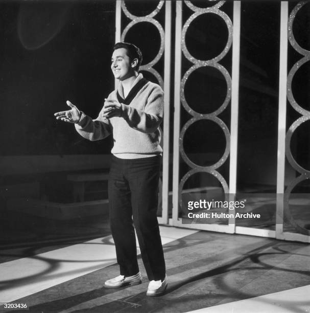 Full-length image of American singer/songwriter Neil Sedaka, wearing a V-neck sweater and penny loafers, gesturing with his hands while standing on a...