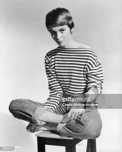 Promotional portrait of American actor Jean Seberg sitting barefoot and cross-legged on a stool, wearing rolled blue jeans and a French-striped...