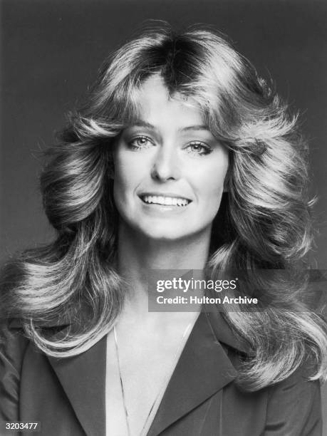 Studio headshot portrait of American actor Farrah Fawcett smiling while wearing a dark blouse and a chain. Fawcett has her trademark layered, wavy...