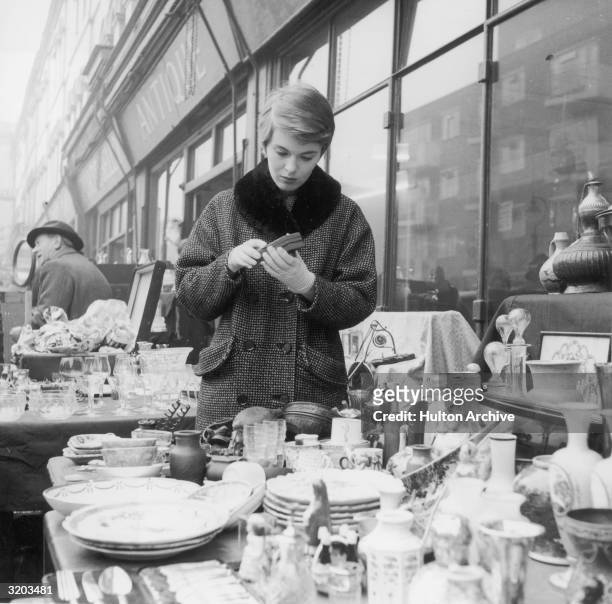 American actor Jean Seberg wears a double-breasted checked overcoat with a fur collar while examining items at a flea market, Paris.