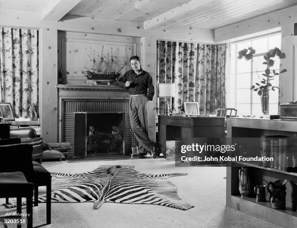 Tasmanian film star Errol Flynn leans nonchalantly against a mantelpiece. A model ship stands on the shelf beside him, and a zebra skin rug lies on...